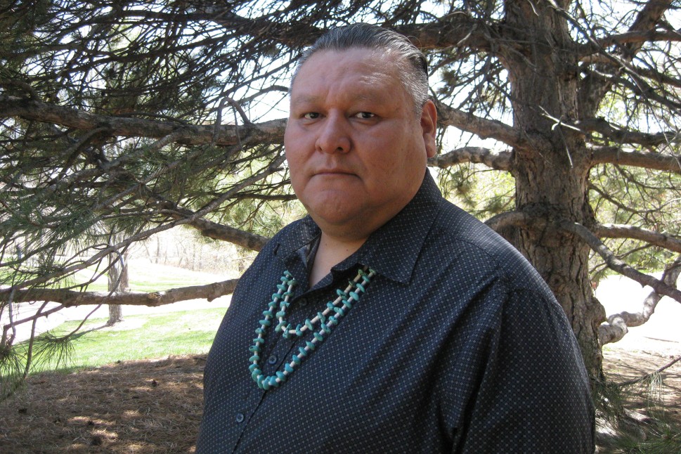 Author standing in front of a pine tree in a black shirt and turquoise necklace. The background is green.